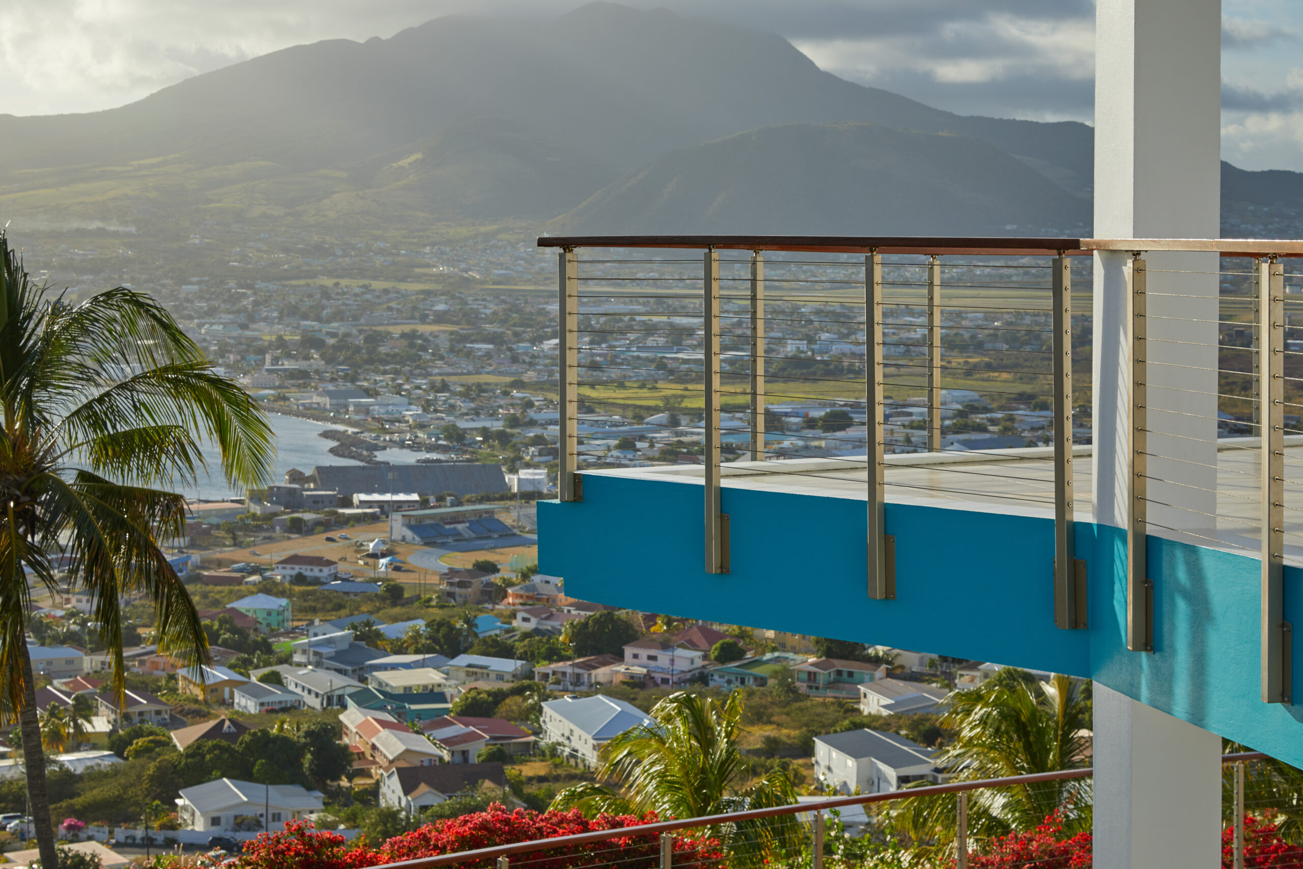 This light blue deck with metal and wire railing overlooks mountains and a valley. The sunlight is diffused behind a cloudy sky. 