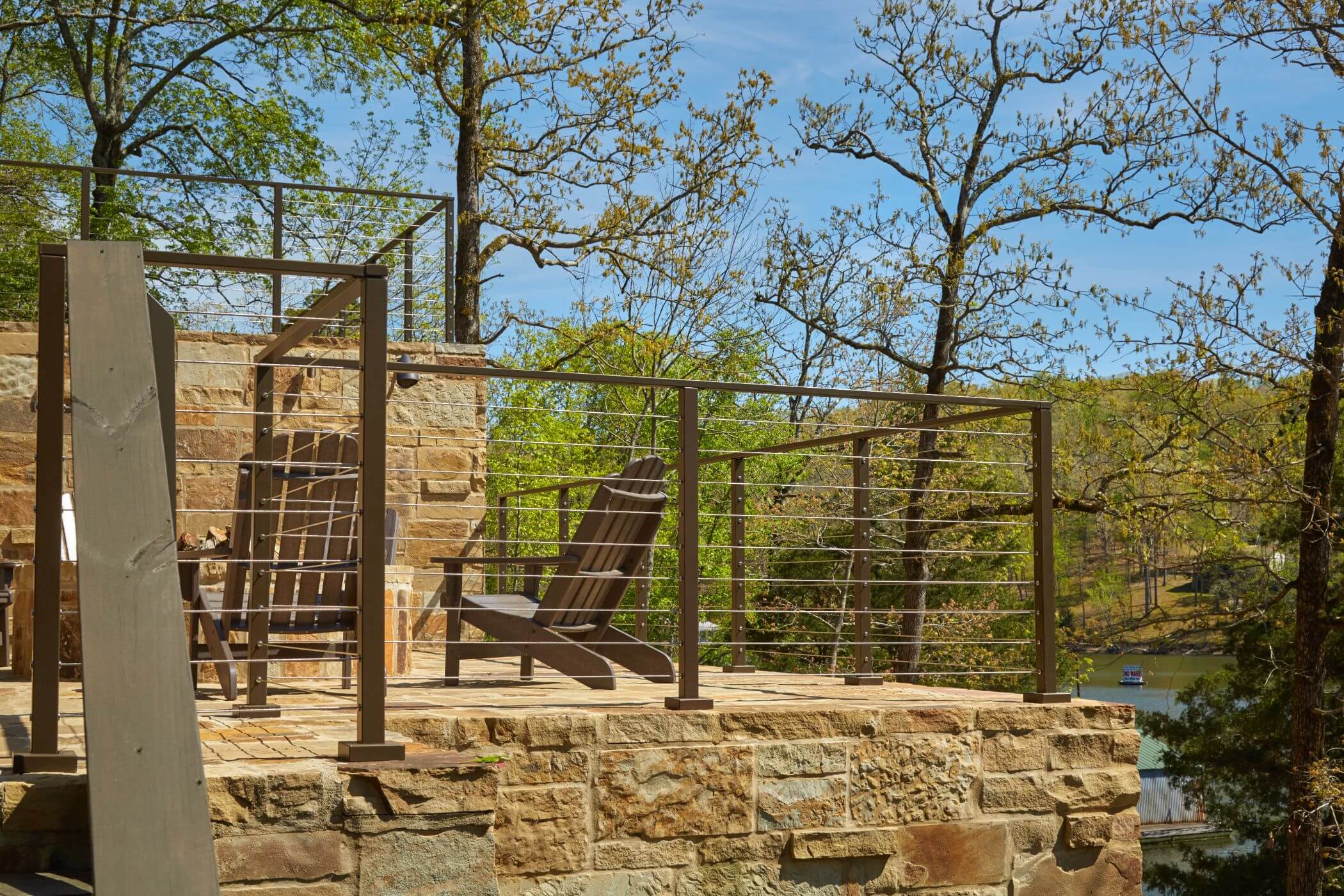 This elevated two-tiered stone platform with metal and wire railing. Two brown chairs sit on the platform. Trees and a body of water are in the background. 