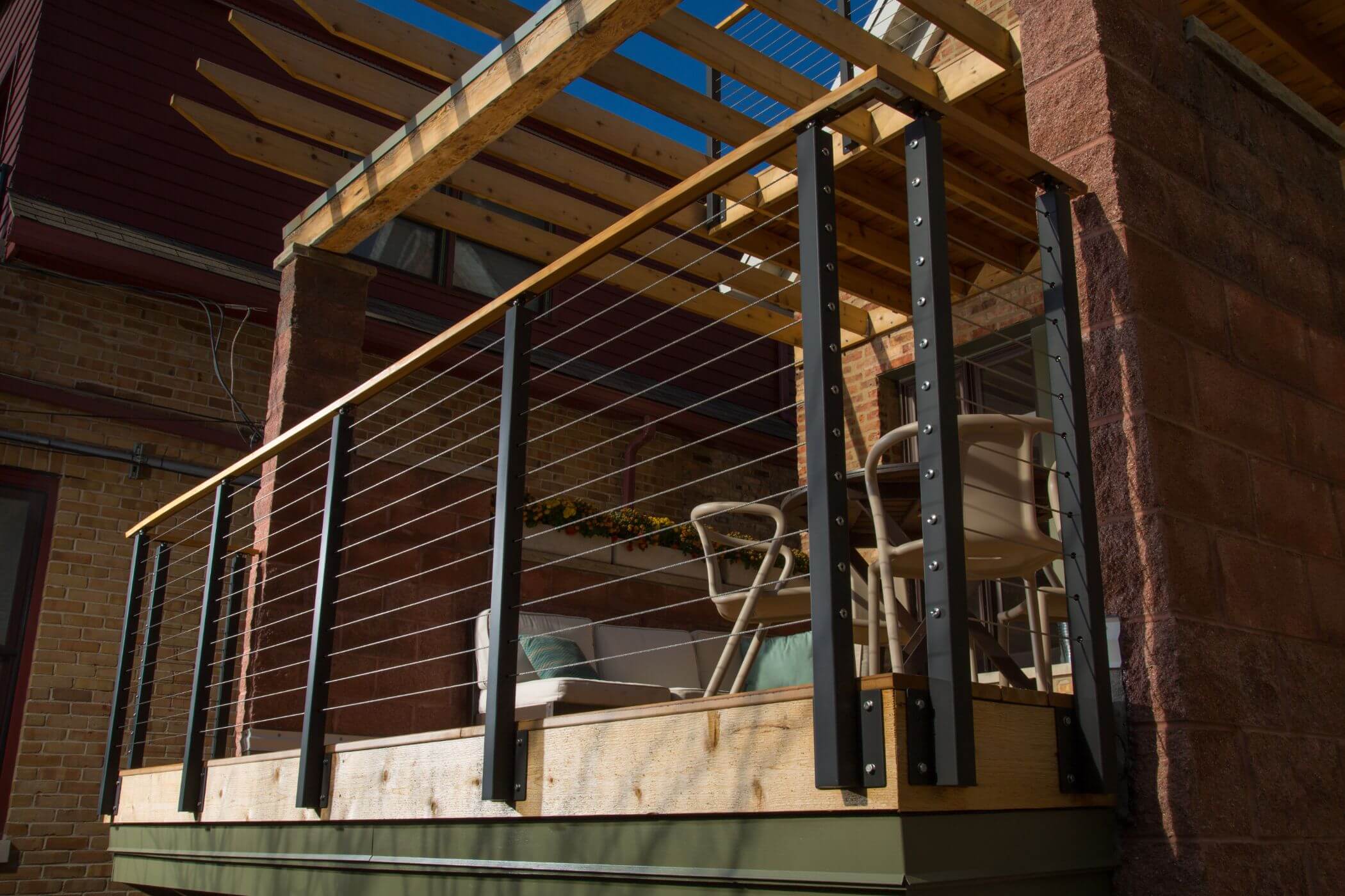 A table with chairs and a couch sits upon a wooden deck with metal wiring and black railings. A wooden gazebo and additional deck hang above.