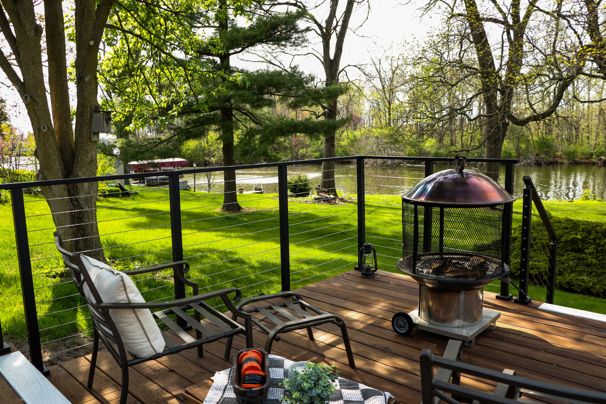 This wooden deck with a fire pit and black chair overlooks the yard with trees and a body of water. 