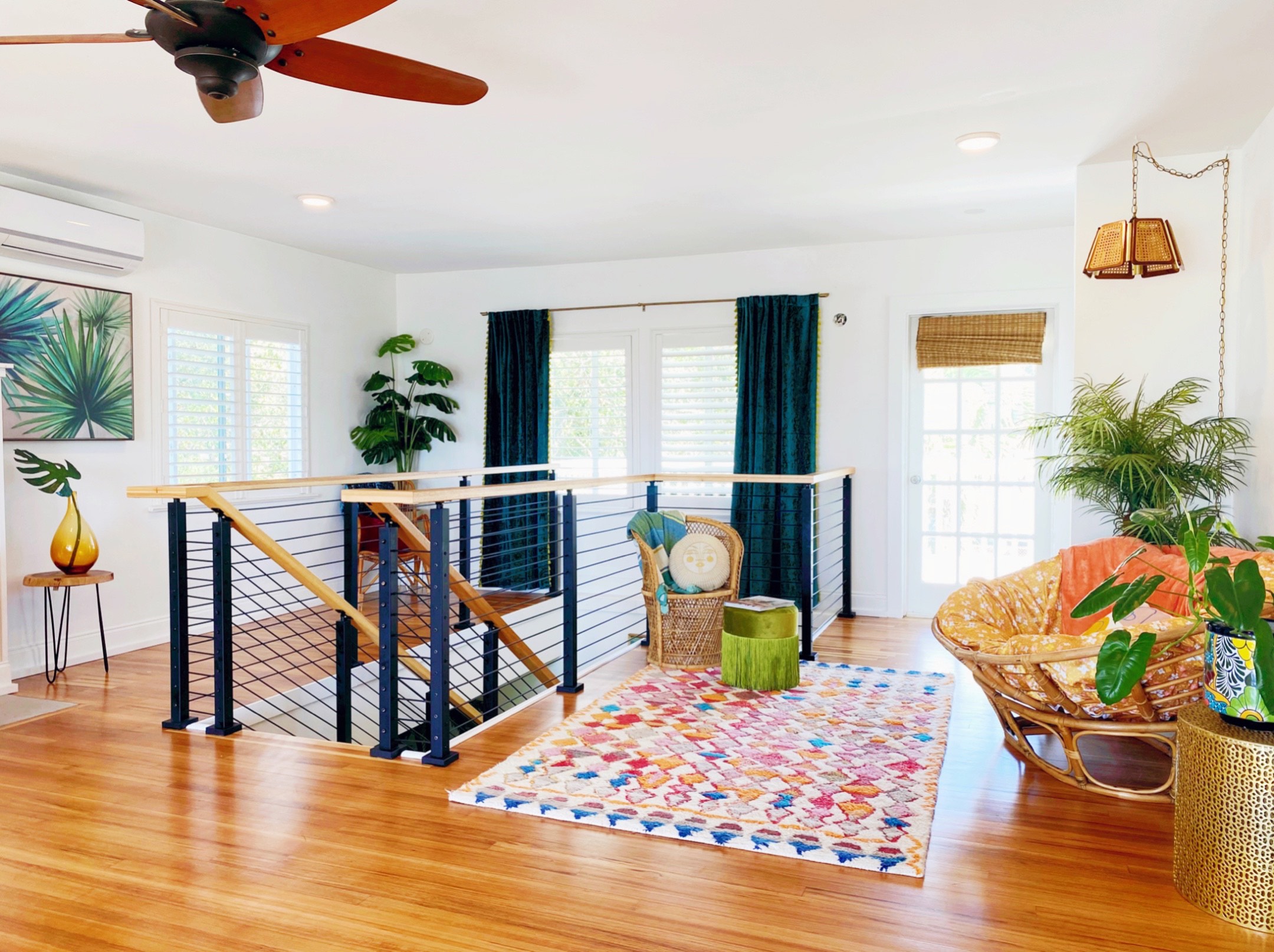 This upstairs space has wooden flooring with a wooden ceiling fan. There is a multi-colored patterned rug on the floor with a wicker chair and ottoman in the corner with a pendant lamp hanging above a bigger chair. 