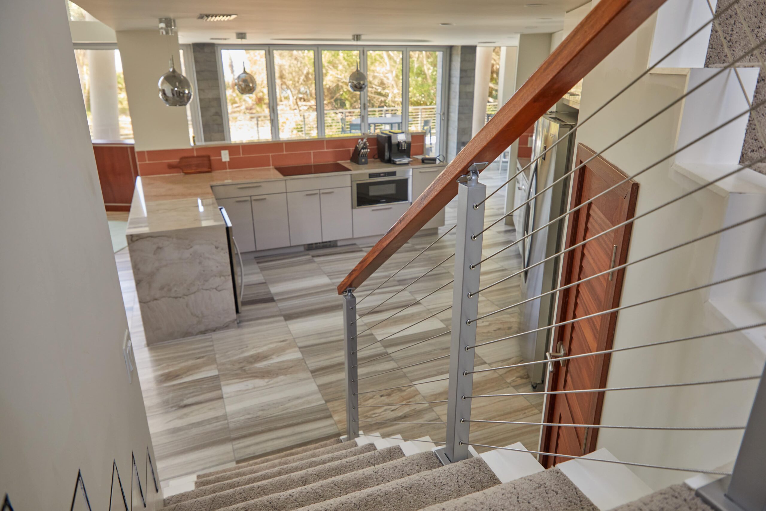 This house has steps leading down to the kitchen. The flooring is small planks of light-colored wood. The kitchen has a marble counter with white cabinets and silver appliances.