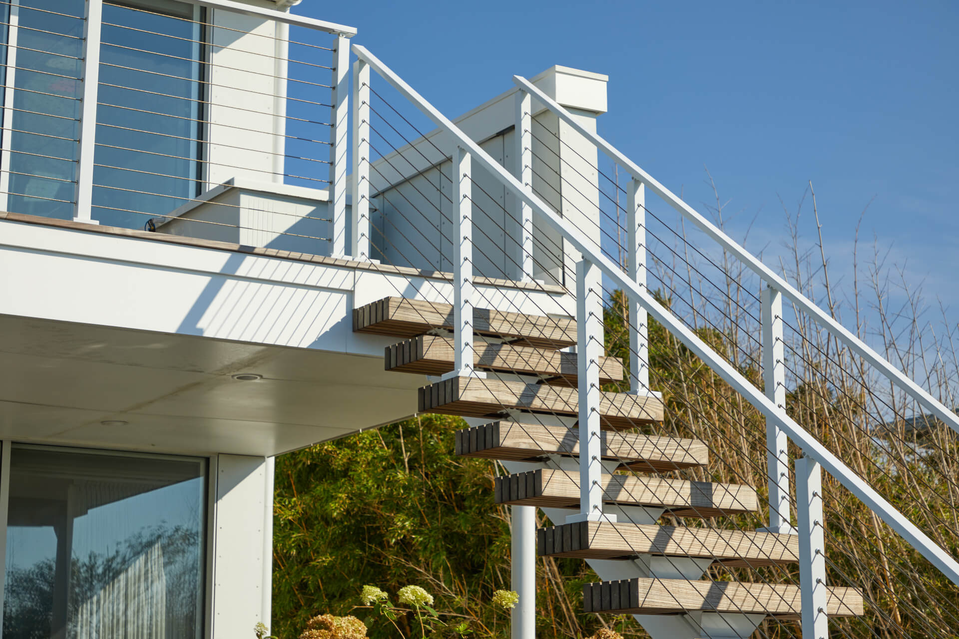 This staircase leads to the balcony of a white house. The staircase has dark wooden steps with floating treads and a metal handrail. There are green plants and a blue sky in the background.